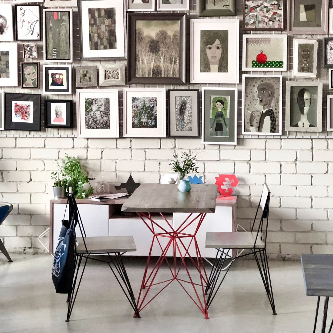 A styled dining area featuring a square table with a gray top and a red geometric metal base, surrounded by two wire-frame chairs. The background is a white brick wall covered in a dense gallery wall of framed art.