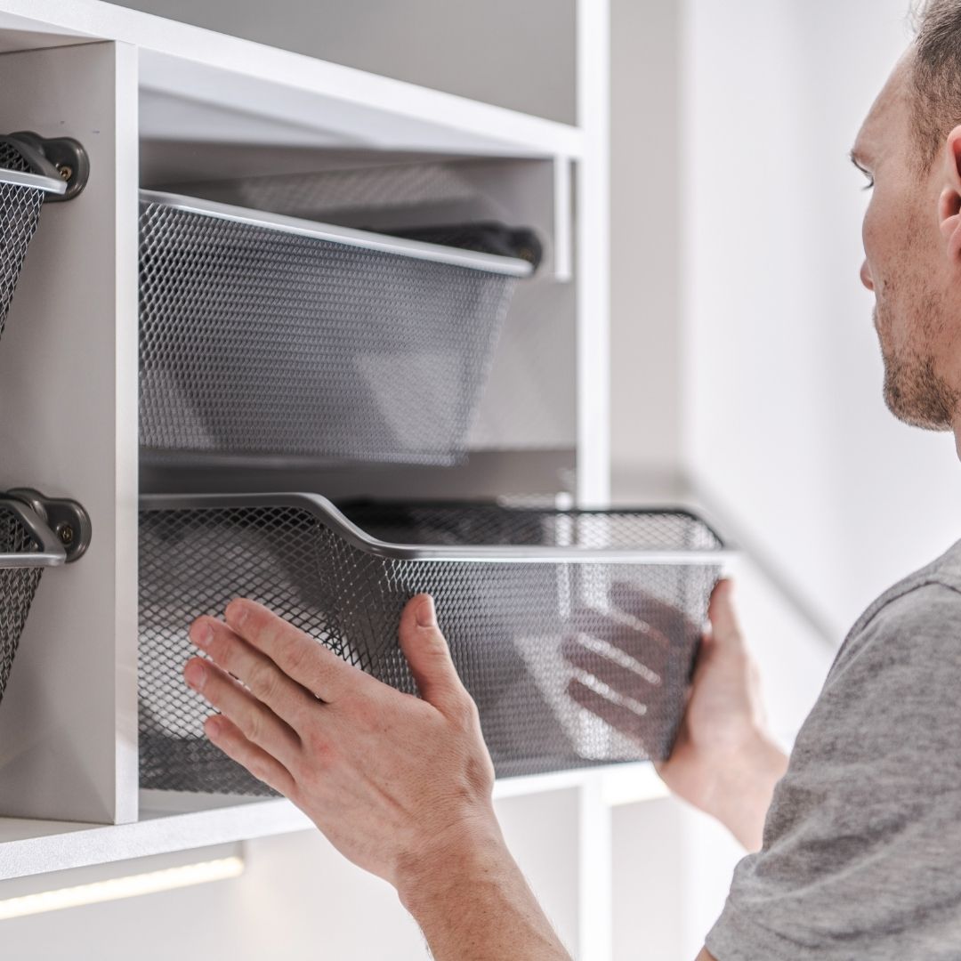 A man pulls out a gray mesh drawer from a white cabinet organizer, with additional drawers visible in the cabinet above.