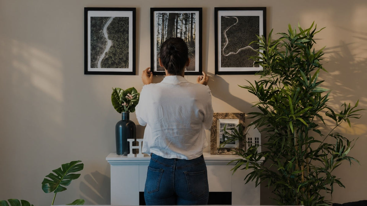 A person viewed from behind hangs three framed black-and-white landscape art prints on a beige wall above a white mantel in a styled room with plants and home decor.